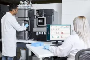 one scientist standing using an instrument, another female scientist at desk using computer data program