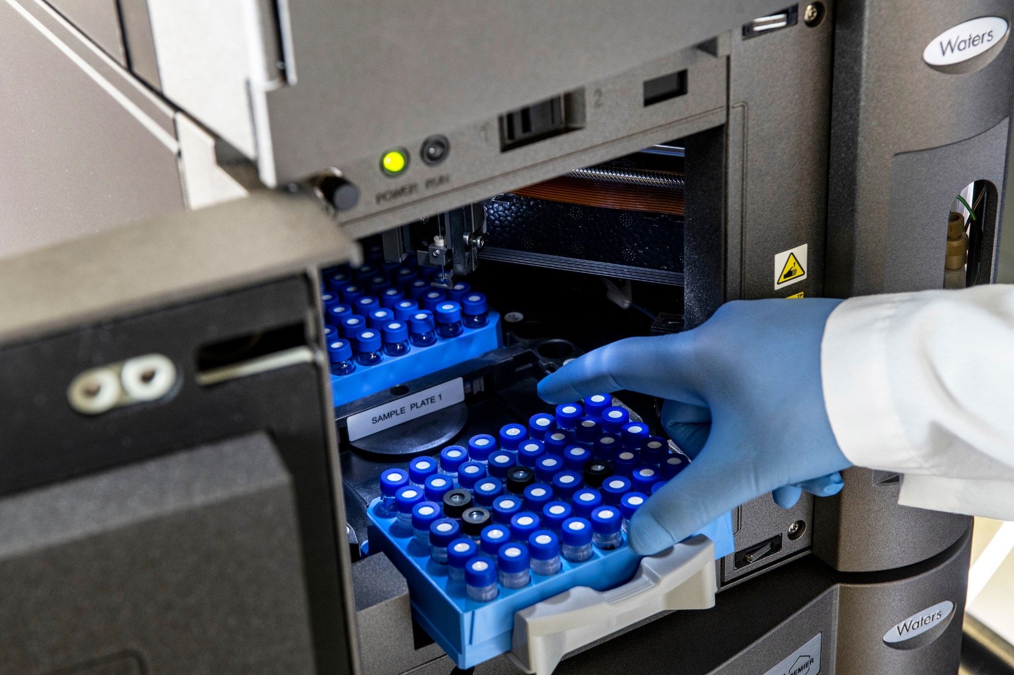 Gloved hand reaching towards a sample tray with blue-capped vials inside the open door of an ACQUITY Premier