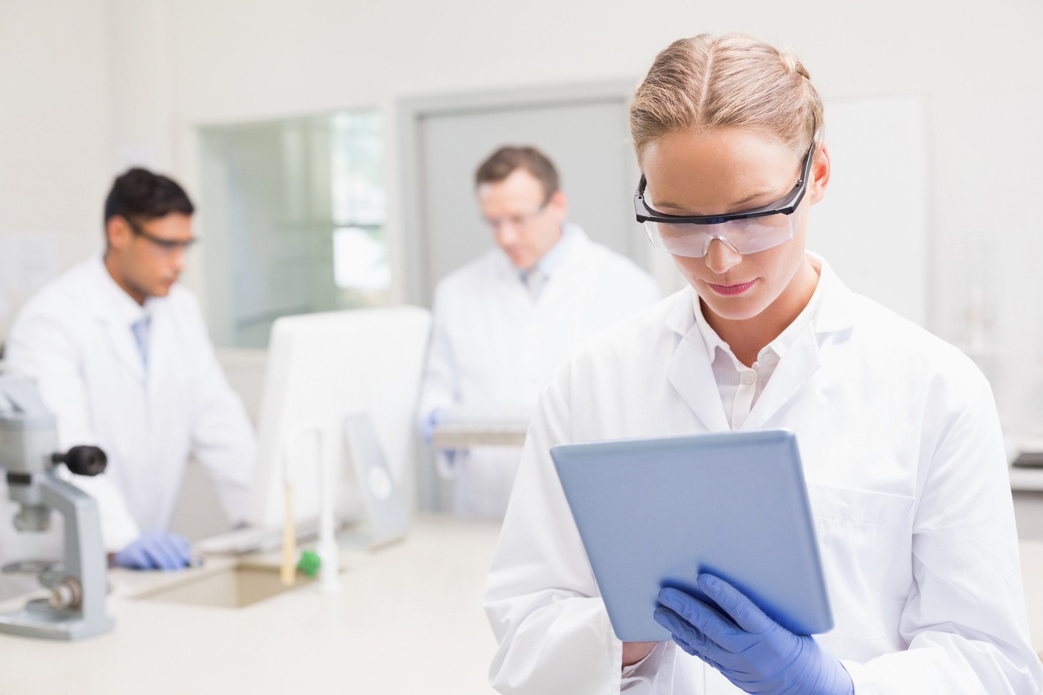 Femme scientifique portant des lunettes et utilisant une tablette pendant que des collègues travaillent derrière elle